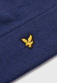 Tissu tricot marine présentant un logo brodé jaune et noir d'une créature ailée dans le coin inférieur gauche. La texture est légèrement texturée.