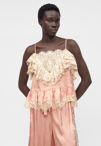 Γυναίκα που φοράει ροζ σατέν σετ με καμισόλ και παντελόνι με κρεμ δαντελένιες βολάν και διαφανή καρδιά στο πάνω μέρος, στέκεται σε απλό φόντο.