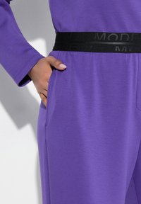 Lila Stoffkleidung mit langen Ärmeln, ausgestattet mit einem schwarzen elastischen Taillenbund mit dem Text "MODEL". Enthält seitliche Taschen für Funktionalität.