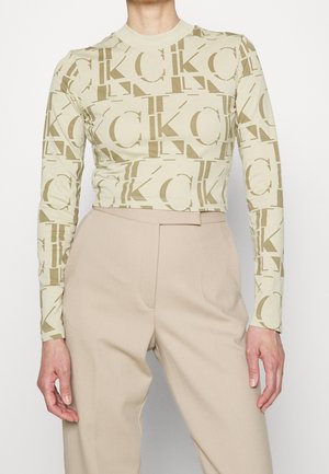 Personne portant un pantalon beige taille haute et un top ajusté à manches longues avec un grand motif "CK" beige et brun clair.