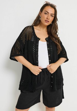 Kvinde iført sort hæklet cardigan med knapper over hvid top og sorte shorts, justerer taljebåndet, med solbriller på hovedet og langt bølget hår.