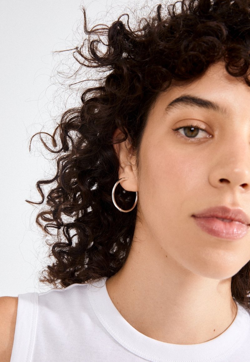 Pendiente de aro plateado en una modelo con cabello rizado y una blusa blanca sin mangas. Enfoque en primer plano en el diseño circular del pendiente y su textura brillante.