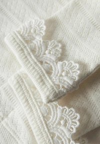 Witte gebreide stof met een gestructureerd patroon en kanten randje aan de randen. Het kant heeft ingewikkelde bloemmotieven.