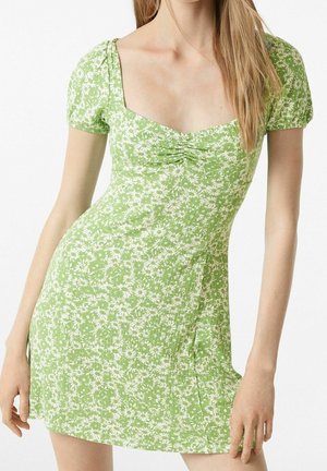Femme en robe verte à manches courtes avec motif floral blanc et centre froncé, debout devant un fond blanc uni.