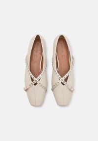 Zapatos de cuero beige con punta cuadrada, presentando correas entrelazadas adornadas con pequeñas tachuelas decorativas a lo largo de los bordes.