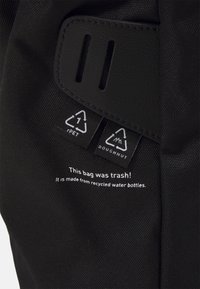 Sac en bouteille d'eau recyclée noir avec texture textile, étiquettes portant des symboles de recyclage, et texte indiquant le matériau et la marque.