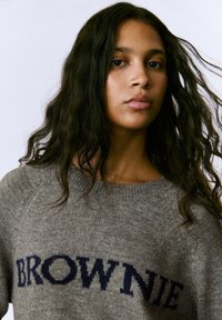 Suéter gris de punto con la palabra "BROWNIE" en letras azul oscuro en el pecho; presenta un cuello redondo y mangas raglán.