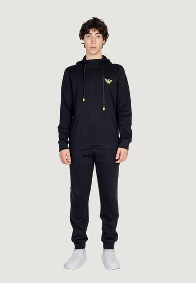 Ensemble sweatshirt à capuche et pantalon de survêtement noir en tissu doux. La capuche est dotée d'une poche avant et de cordons de serrage accentués de jaune. Baskets blanches.