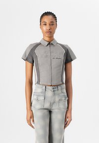 Polo grigio tagliato con maniche corte, chiusura a zip, logo ricamato "GUESS ORIGINALS", abbinato a jeans chiari strappati.