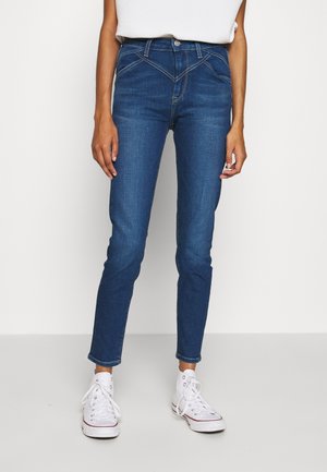 Osoba ubrana w dopasowane niebieskie dżinsy z detalem w kształcie litery V w talii oraz białe wysokie trampki, stojąca na jednolitym tle.