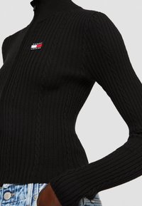 Lähikuva henkilöstä, joka pitelee mustaa ribbikudottua neuletta, jossa on pieni Tommy Jeans -logo, sekä vaaleansinisiä farkkuja.