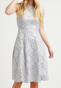 Ärmlös knälång silverfärgad klänning i spets med blommönster, figursydd midja och genomskinlig krage, bärs av en kvinna med blont hår.