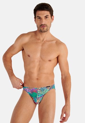 Gespierde man met kort donker haar die kleurrijke zwembroek met tropisch patroon draagt tegen een effen witte achtergrond.