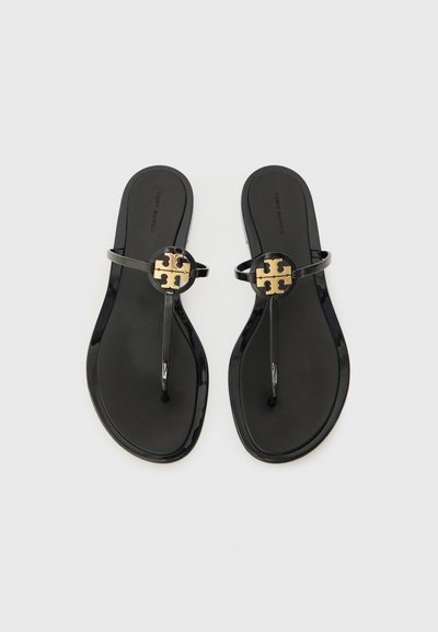 Sorte flip-flops med en blank finish, avsmalnende stropper og en sirkelformet gullakent med et utskåret design foran.
