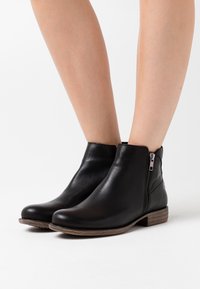 Botas de cuero negro hasta el tobillo con punta redondeada, cremallera lateral y tacón apilado. Textura suave y diseño minimalista.