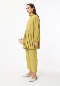 Limegrünes, plissiertes Tunika mit hohem Kragen, kombiniert mit passenden schmalen Hosen. Weiße Mules vervollständigen das Outfit und zeigen ein minimalistisches Design.