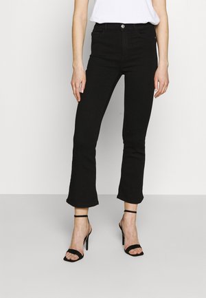 Kobieta ubrana w czarne, skrócone jeansy z rozszerzanymi nogawkami, czarne szpilki na kostkę i biały top, stojąca na jednolitym tle.