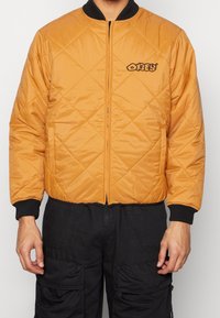 Veste bomber matelassée en jaune moutarde avec des poignets côtelés noirs, des poches avant et un logo "OBEY" brodé sur la poitrine gauche.