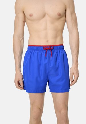 Bañador azul para hombre con cintura roja y cordón, usado por una persona sin camiseta desde el pecho medio hasta el muslo medio, de pie contra un fondo liso.