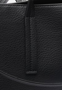 Primo piano di una borsa in pelle nera, con una superficie testurizzata, cuciture in evidenza e un dettaglio di tracolla stretta. Bordi lisci e forma arrotondata.