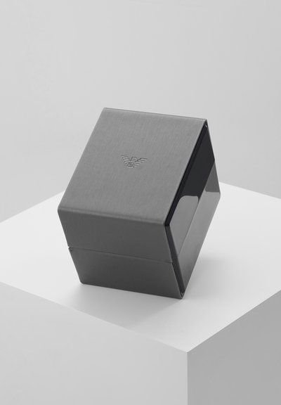 Caja angulada cerrada de color gris y negro con logo en relieve en la parte superior, colocada sobre un pedestal blanco contra un fondo gris claro.