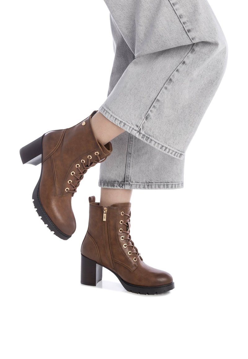 Botas de cuero marrón hasta el tobillo con un tacón grueso, cordones en la parte delantera, suela texturizada y detalle de cremallera lateral. Llevadas con jeans amplios grises.