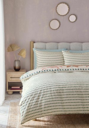 Bett mit hellblau-weiß gestreifter Bettwäsche, gepolstertem Kopfteil, hölzernem Nachttisch mit Vase und getrockneten Palmblättern und drei runden Wandspiegeln.