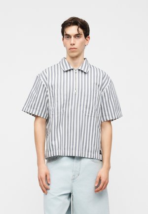 Tânăr care poartă o cămașă albă largă cu mâneci scurte, cu dungi negre verticale și blugi denim bleu deschis, stând în picioare pe un fundal simplu.