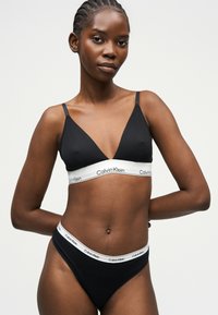 Γυναίκα με πλεγμένα μαλλιά που φοράει μαύρο bralette Calvin Klein και μαύρο σλιπ, μπροστά σε μια απλή ανοιχτόχρωμη επιφάνεια.