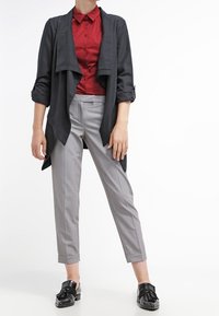 Blazer noir drapé sur une chemise rouge à col, associée à un pantalon fuselé gris clair et des mocassins noirs à franges.