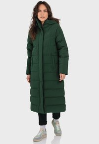 Langer grüner Puffermantel mit Kapuze, der ein gestepptes Design und seitliche Taschen aufweist. Getragen über schwarzen Hosen und mehrfarbigen Sneakers.