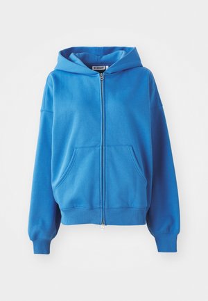 Zila zip-up kapuce ar kapuci, divām priekšējām kabatām, ribotām piedurknēm un apakšmalu. Izgatavota no mīksta auduma, ar gludu tekstūru un vienkāršu dizainu.