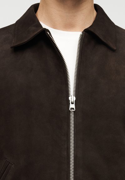 Καφέ σατινέ μπουφάν με μυτερό γιακά, που διαθέτει ασημένιο φερμουάρ και δύο πλαϊνές τσέπες, συνδυασμένο με λευκό μπλουζάκι με στρογγυλή λαιμόκοψη από κάτω.