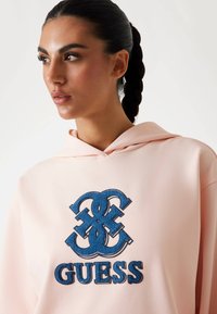 Sudadera rosa con un gran logo en denim azul de "GUESS" y letras entrelazadas. Tejido suave, corte relajado y detalle de capucha con cordones.