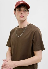 Bruine katoenen t-shirt met korte mouwen, ronde halslijn, gecombineerd met een kastanjebruine pet met witte tekst. Een zilveren kettinghalsketting accentueert de look.