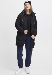 Svart polstret jakke med oransje snorer, båret over en marineblå genser og joggebukser. Hvite treningssko med teksturerte såler fullfører antrekket.