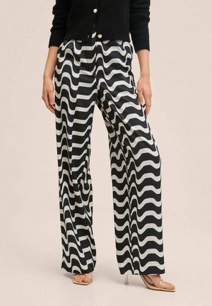 Femme portant un pantalon à jambes larges avec un motif ondulé noir et blanc, des talons à brides transparentes et un cardigan noir boutonné.