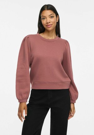 Kvinde med langt mørkt hår iført en malva puff-ærmet sweatshirt og sorte bukser, stående mod en ensfarvet lys baggrund.