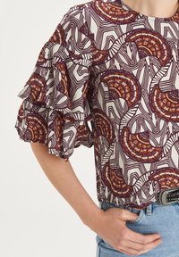 Haut à manches courtes en tissu léger, avec une base blanche ornée de motifs en éventail violets, orange et marron, des manches à volants et une coupe décontractée.