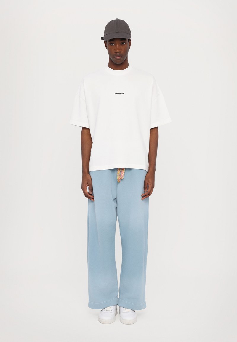 T-shirt blanche ample avec le texte "Bonsai", associé à un pantalon large bleu clair et des baskets blanches. Un homme porte une casquette grise.