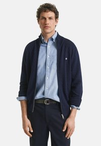Chaqueta de navy con cremallera y patrón de punto texturizado, llevada sobre una camisa de botones azul claro. Modelo estilizado con un cinturón negro y pantalones navy.