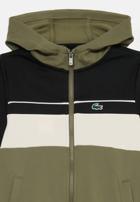 Veste à capuche zippée avec des blocs de couleur olive, noir et blanc, poches avant et un petit logo de crocodile vert sur la poitrine.