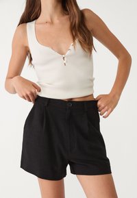 Femme ajustant un short noir taille haute avec un bouton, portant un crop top sans manches blanc à côtes avec des boutons, devant un fond uni.