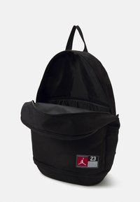 Zaino nero realizzato in tessuto resistente, dotato di un ampio comparto principale e una tasca anteriore con zip, decorata con un patch logo rosso e grigio.