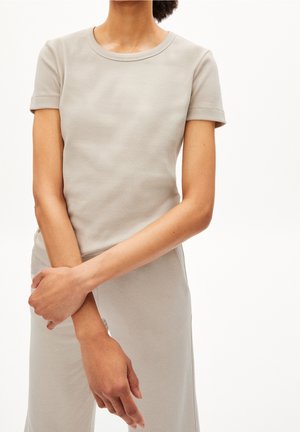 Personne portant une chemise côtelée beige à manches courtes et un pantalon assorti, debout les bras croisés devant un fond blanc.