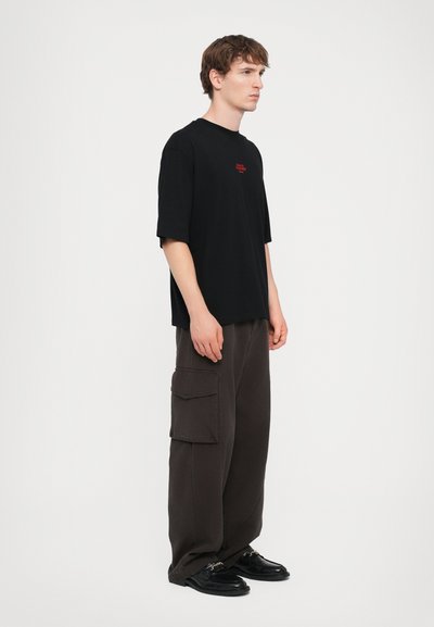 T-shirt noir à manches courtes avec texte rouge, associé à un pantalon cargo brun ample avec poches latérales et chaussures noires.