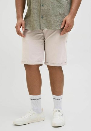 Uomo con pantaloncini beige risvoltati, camicia verde oliva con bottoni, calzini bianchi del marchio Jack & Jones e scarpe da ginnastica bianche, in piedi su uno sfondo semplice.
