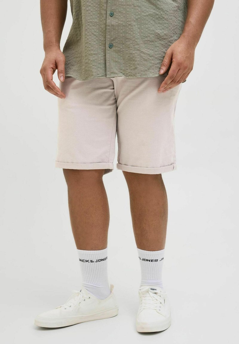 Homme portant un short beige retroussé, une chemise vert olive boutonnée, des chaussettes blanches de marque Jack & Jones et des baskets blanches, debout sur un fond uni.