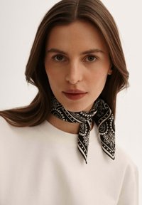 Czarny szalik w wzory wykonany z lekkiego materiału, zdobiony wzorami paisley. Związany wokół szyi, doskonale komponujący się z gładką białą bluzką.