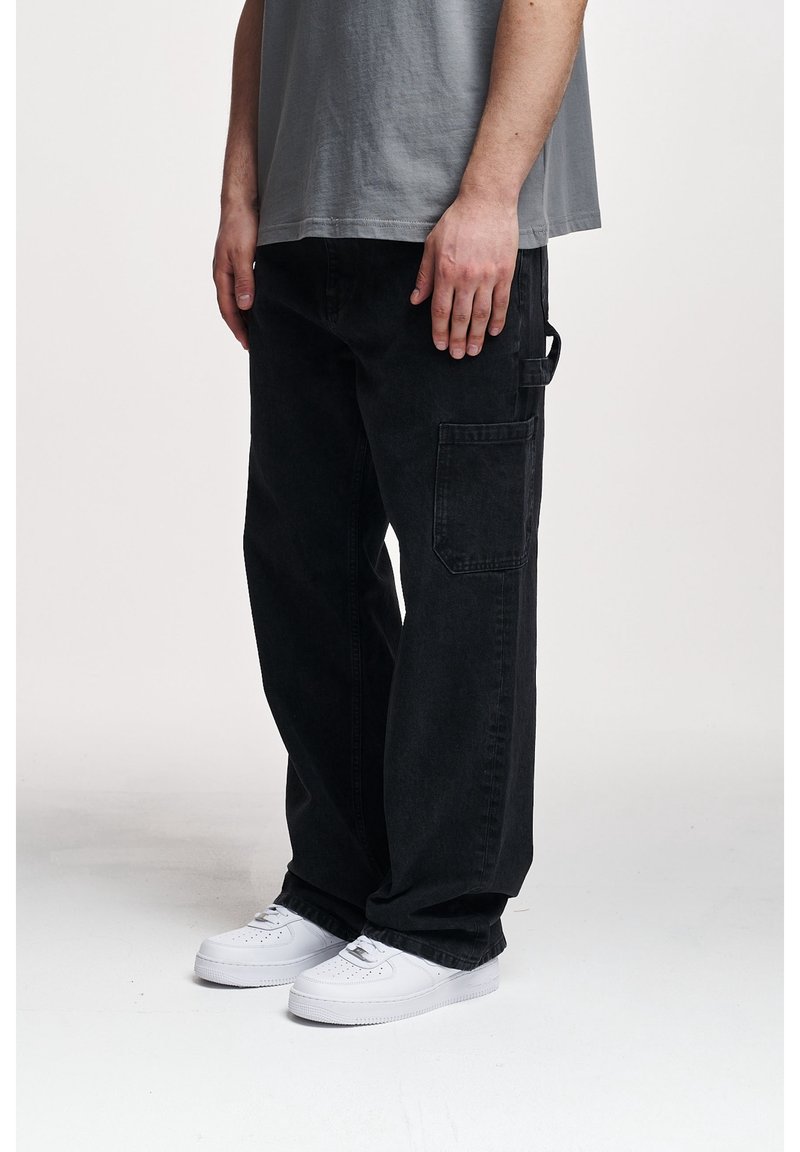 Personne portant un pantalon cargo noir ample et des baskets blanches, debout avec les mains détendues le long du corps sur un fond uni.